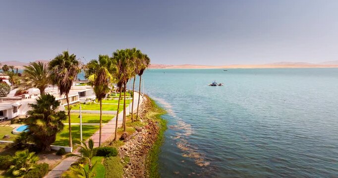 Palm-lined waterfront homes beside calm blue water and desert dunes in Paracas, Peru, drone dolly above resort in early morning
