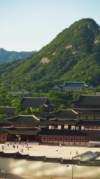 Tourists crossing sunlit Gyeongbokgung Palace courtyard with tiled roofs, Cheong Wa Dae and forested Bugaksan mountain rising behind in central Seoul on clear summer day - aeria vertical shot