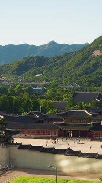 Aerial vertical shot of tourists strolling across sunlit Gyeongbokgung Palace courtyard with Heungnyemun gate, Cheong Wa Dae and green Bukhansan ridge rising behind under clear summer sky