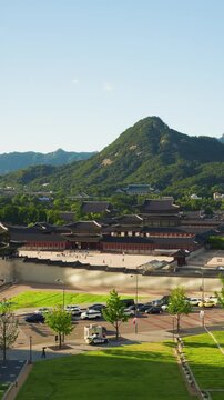 Wide elevated view of Gyeongbokgung Palace rooftops, Cheong Wa Dae and forested Bugaksan mountain above Gwanghwamun Square lawn with cars and police vehicle passing on sunny summer day