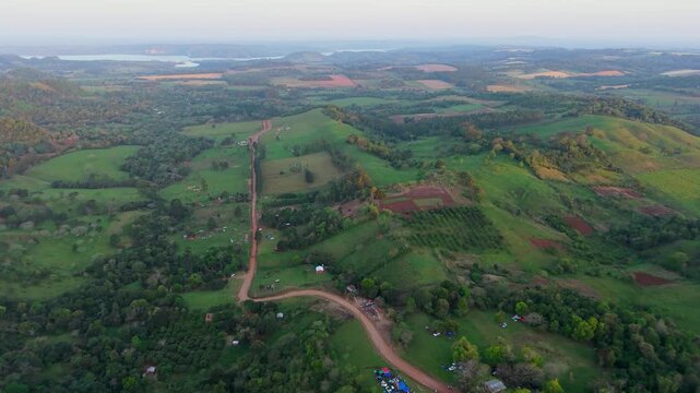 Gliding over red dirt rally tracks, green hills, and surrounding vegetation under late afternoon sky before sunset in Nueva Alborada, Paraguay.