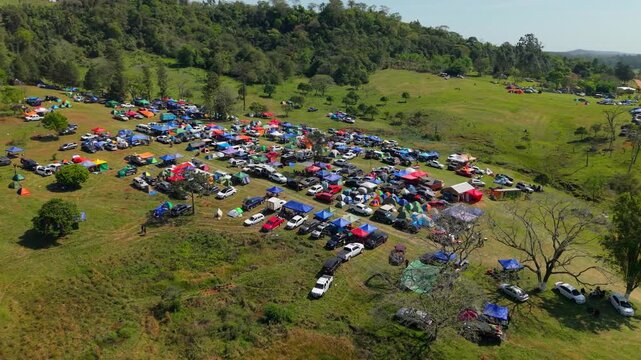 Orbiting left above crowded campsite area packed with vehicles and tents next to WRC Rally race tracks surrounded by lush green terrain on sunny day.