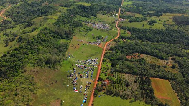Descending drone approach tilted down over red dirt race tracks, green vegetation, and crowded spectator campsites during a sunny day at WRC Rally 2026. 4K at 24 fps.
