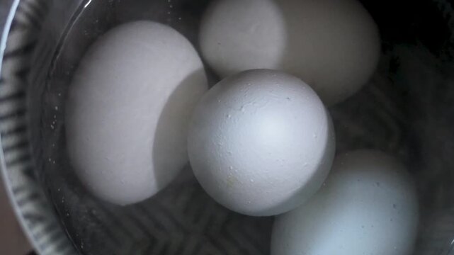 Smooth close-up shot of white eggs gently boiling in a metal pot of water, bubbles rising softly. Natural lighting highlights texture and detail, capturing a calm, kitchen cooking moment.
