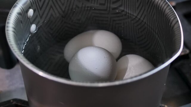 Three eggs sit inside a metal saucepan on a stovetop during food preparation, showing white shells and stainless steel cookware in close up