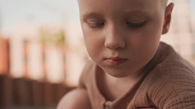 Young boy sits outdoors focused on small yellow object in his hands. Boy wears brown textured shirt as boy examines item closely against urban background with blurred buildings and soft