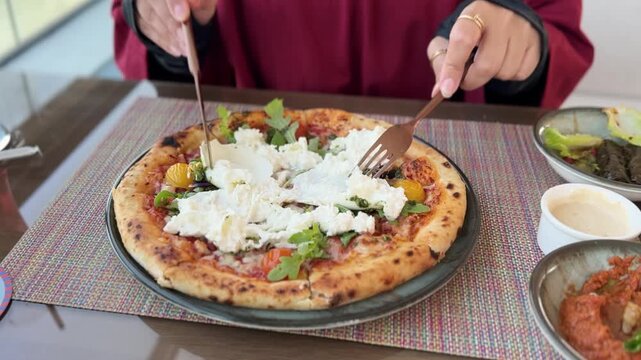 Close-up of hands using a knife and fork to cut a fresh burrata pizza with tomatoes and arugula. A person enjoys a daytime lunch at a restaurant table with side dishes.