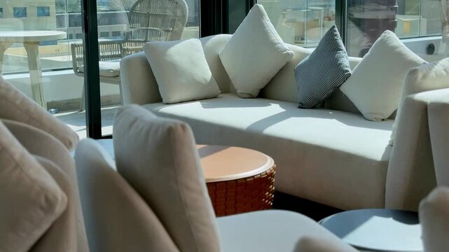 A modern white sofa with decorative pillows and a round coffee table in a sunlit indoor lounge. Large glass windows reveal an outdoor patio and urban city background during bright daylight.