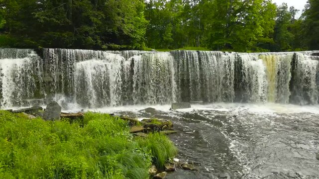 Beautiful aerial footage flying towards the majestic Keila Joa waterfall in Estonia during a bright summer day. The powerful water cascades over the rocky ledge into the river below