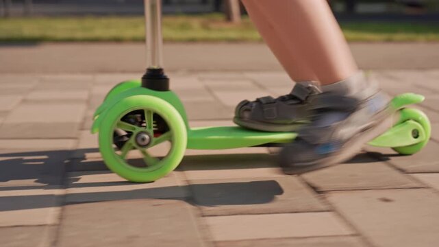 Young boy rides green kick scooter on paved ground with another child visible in background. scene features outdoor daylight and clear weather details like shadows from surrounding objects