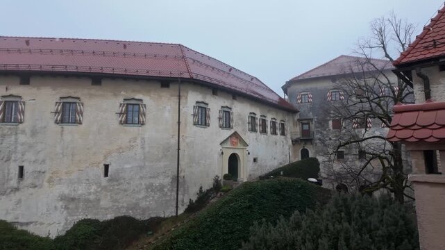 Bled Castle in Slovenia with medieval stone walls, arched windows and an inner courtyard, surrounded by bare trees in overcast winter weather, panoramic exterior view.