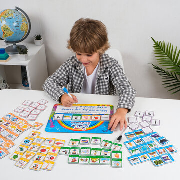Schoolboy completes an English sentence writing worksheet at a table surrounded by colorful grammar and picture cards, practicing handwriting, vocabulary and language skills.