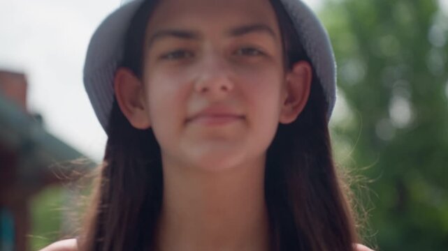Young woman with long dark hair wearing light blue bucket hat stands outdoors in bright daylight. background features green foliage and parts of outdoor structure suggesting casual