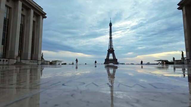Low angle view across the wet Trocad&eacute;ro plaza toward the Eiffel Tower at blue hour, with a few distant pedestrians crossing the open square.