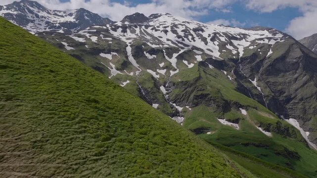 Lush Na&szlig;feld hillside pulls past camera to reveal striped summer snowfields and thin waterfalls on the opposite range.