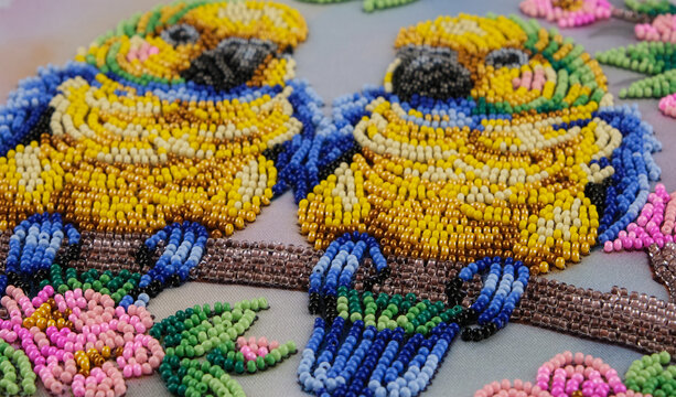 Handmade beadwork features a pair of colorful birds sitting side by side on a branch with delicate pink flowers, creating a decorative scene of nature, harmony and togetherness.