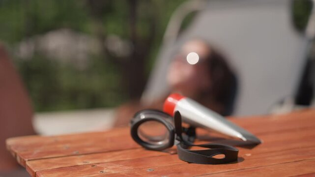 Woman relaxes outdoors with swimming goggles and tube of sunscreen on wooden table. scene features soft natural light greenery in background and minimal camera movement to emphasize still