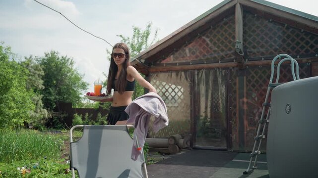 Young woman in black swimsuit carries plate with fruit and orange drink placing it on wooden table near outdoor furniture. Woman holds towel while walking through garden setting