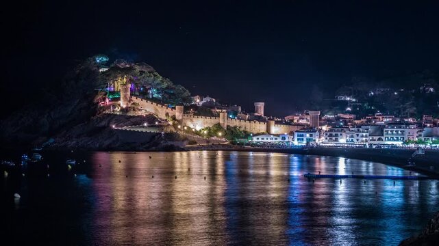 Panoramic night view of the illuminated medieval castle and old town of Tossa de Mar on the Costa Brava, with colorful lights reflecting on the calm mediterranean sea water in Catalonia, Spain