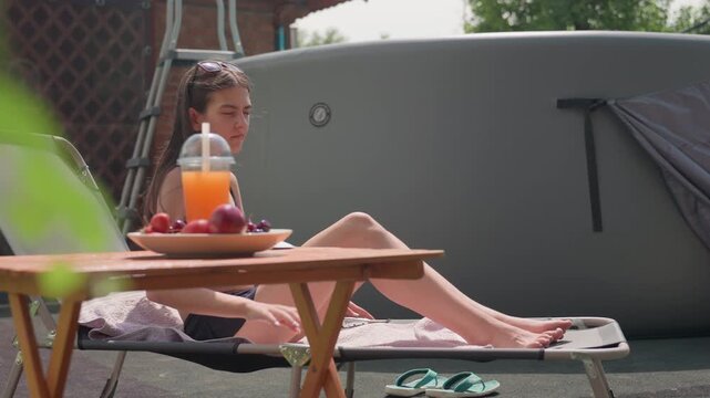 Young woman relaxes on sun lounger outdoors adjusting her posture and enjoying setting. wooden table with fruit bowl and drink is nearby along with flip flops placed beside them. scene