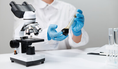 Laboratory oil sample testing setup with microscope and gloved hands holding dark flask for...
