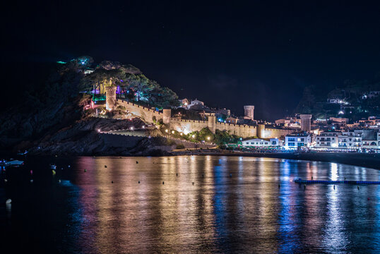 Panoramic night view of the illuminated medieval castle and old town of Tossa de Mar on the Costa Brava, with colorful lights reflecting on the calm mediterranean sea water in Catalonia, Spain
