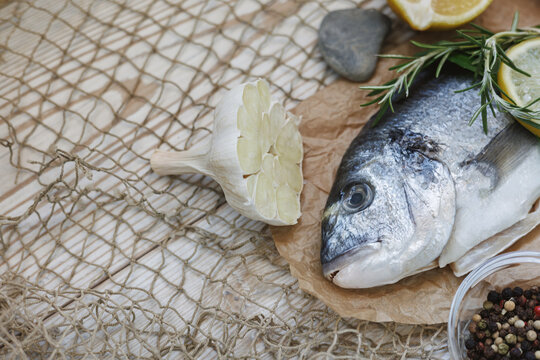 Fresh gilthead sea bream with garlic rosemary lemon and mixed pepper on kraft paper over fishing net, rustic Mediterranean seafood cooking concept with copy space