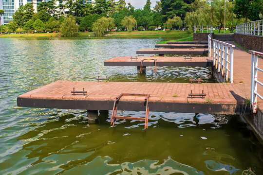 Stone paved walkway with piers docks and boat cleats along lake shore in city park with green trees, railing and buildings background