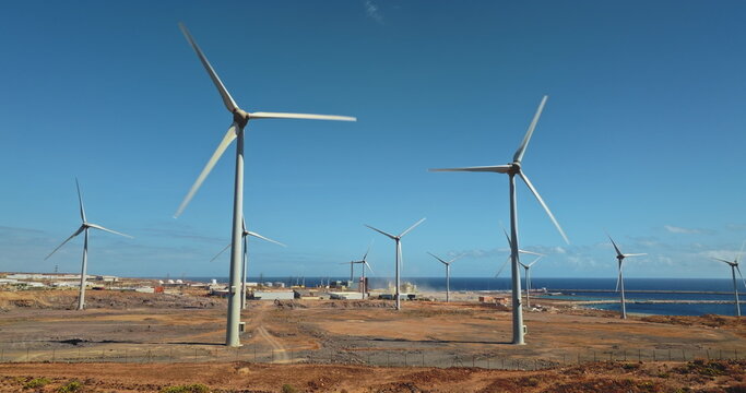 Wind turbines spinning blades on arid land near the ocean in Tenerife against blue sky, representing sustainable renewable energy and a commitment to environmental protection. Aerial drone panorama