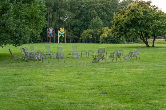 Empty metal mesh lounge chairs are arranged in a loose circle on a green park lawn, with colorful playground figures visible behind trees.