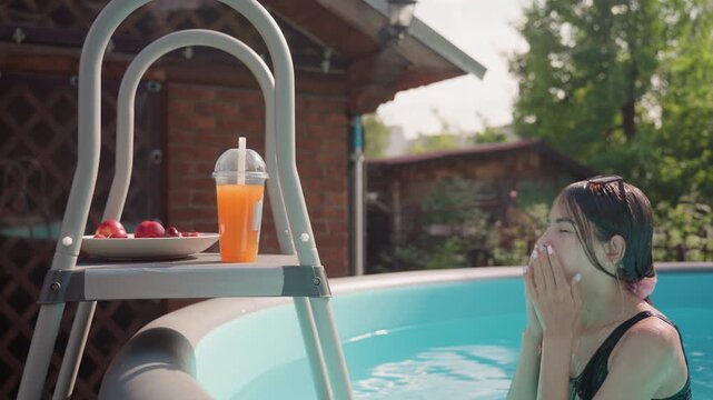 Woman in pool swimsuit enjoys time by outdoor swimming pool with fresh fruit and orange juice on ladder. scene features clear blue water green foliage background brick wall structure soft