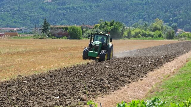 Tractor plowing a harvested wheat field in rural Liguria, Italy, turning dry stubble into dark soil with mountains and countryside in the background. Agriculture concept, agribusiness.