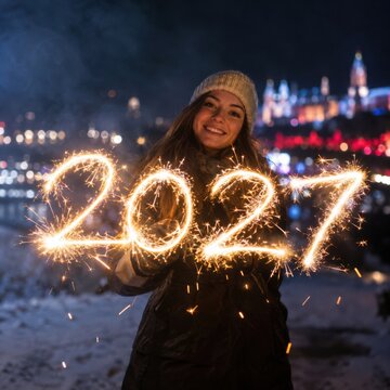Junge Frau begr&uuml;&szlig;t 2027 mit Wunderkerzen &ndash; L&auml;chelnde Frau in winterlicher Kleidung h&auml;lt leuchtende Jahreszahlen vor einer verschneiten Stadt und feiert den Jahreswechsel.