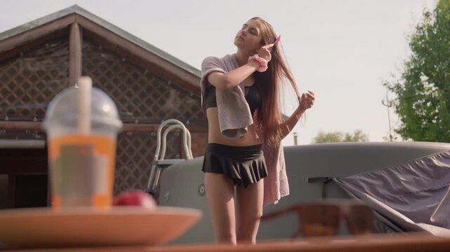 Young woman in black swimsuit and pink towel combs her hair outdoors near above ground pool with sunlight casting soft shadows. plastic cup drink is visible on table nearby while background