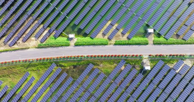 Aerial view of blue solar panels arranged in parallel rows across green ground, divided by a paved road with trees and utility equipment in bright sunlight.