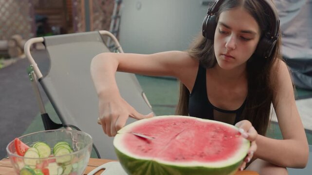 Young woman wearing headphones slices large watermelon with knife while seated at outdoor table. clear bowl containing sliced cucumbers and tomato is visible beside her along with edge