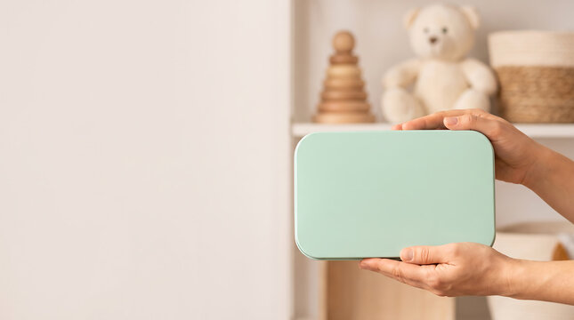 Hands holding blank mint green baby care storage box mockup in nursery room with teddy bear and wooden toys on shelf