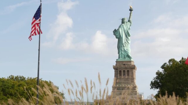 Statue of Liberty on Liberty Island, New York, United States. Cooper monument from behind, democracy and patriotism symbol near Manhattan, NYC USA. American flag waving in wind. Independence concept.