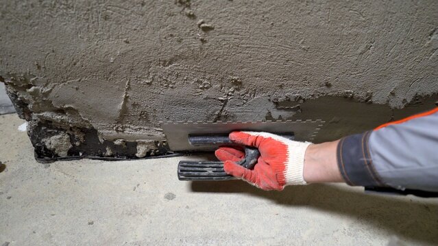 Construction worker in protective glove using notched trowel to apply fresh plaster to rough concrete wall