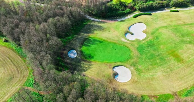 Aerial view of a vibrant green golf course putting area surrounded by sand traps, fairway turf, and adjacent forest trees in bright sunlight.