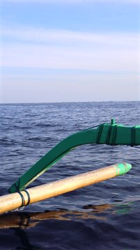 Outrigger canoe moves across ocean water. Curved green outrigger and bamboo float skim along surface while low swells and wide horizon create simple seafaring view with paddle, hull, and wake.