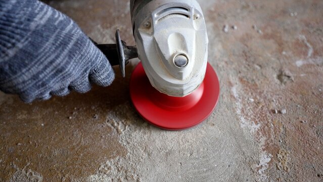 Worker in grey glove using angle grinder with red disc to polish dusty concrete floor during renovation