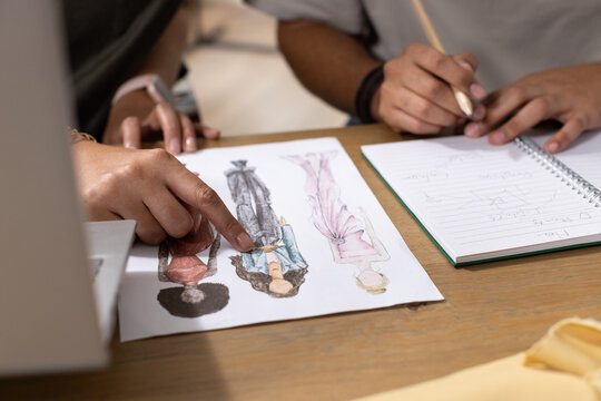 Fashion sketches are being reviewed on wooden table with pointing hand, notebook, pencil, fabric