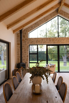 Rustic wooden dining table in vaulted room, featuring ceramic vase with greens, black doors to lawn