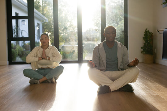 African American couple meditating crossed-legged on hardwood floor near glass doors