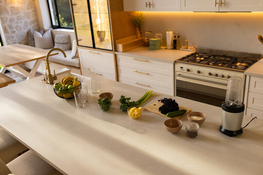 Bright modern kitchen island is showcasing fruit bowl, gold faucet, cutting board with vegetables