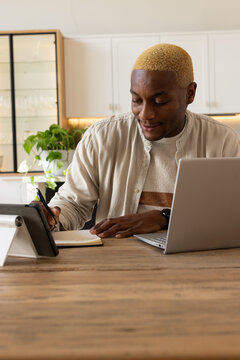 African American man in striped knit writing notes and checking tablet at kitchen table with laptop
