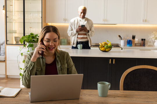 Couple in kitchen with woman at table working on laptop, speaking on smartphone, wearing eyeglasses