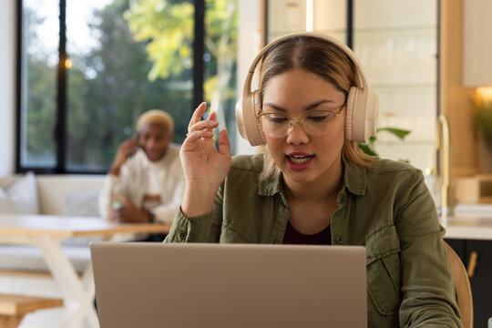 Diverse couple in kitchen, woman speaking on laptop with headset and glasses, partner holding phone