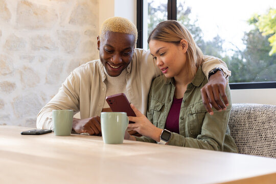 Couple sitting close at light wood dining table in home dining nook, sharing maroon smartphone